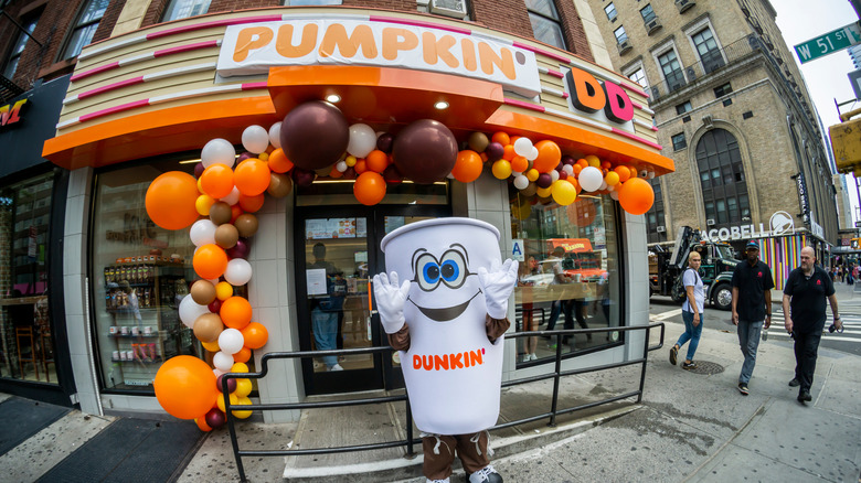 Someone wearing a Dunkin' coffee cup costume outside a Dunkin' store with a pumpkin sign