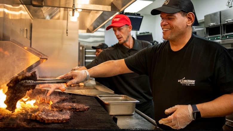 Two LongHorn Steakhouse workers in caps and black shirts make steaks on a grill in an industrial kitchen