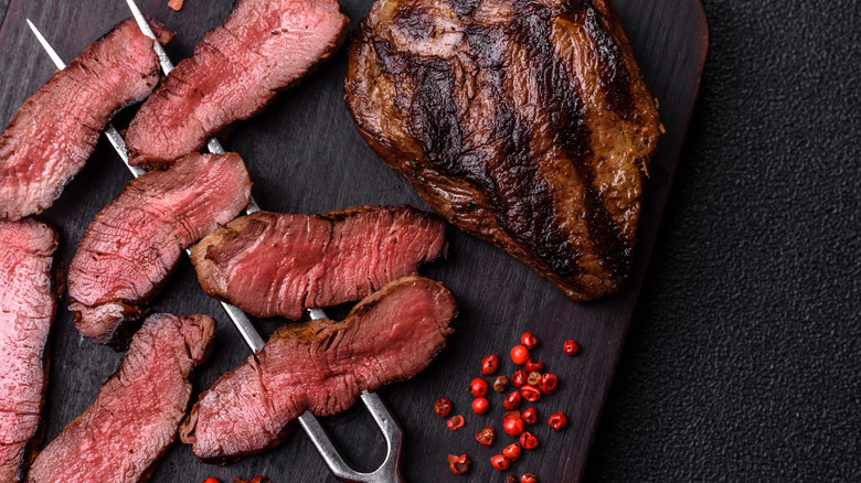 A medium-rare steak, partially sliced, skewered by a long fork, on top of a wooden cutting board