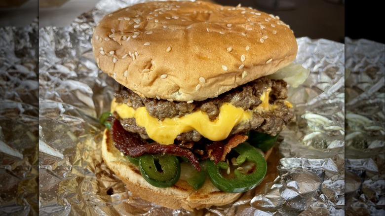 A Five Guys bacon cheeseburger topped with visible jalapenos, sitting on the foil wrapper.