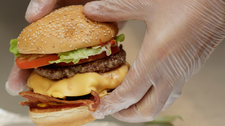 Gloved hands holding a bacon cheeseburger from Five Guys with visible lettuce, tomato, burger patty, cheese, and bacon on a sesame seed bun.