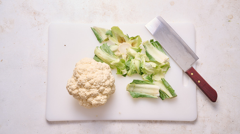 Cauliflower head, stems, leaves, and knife on cutting board