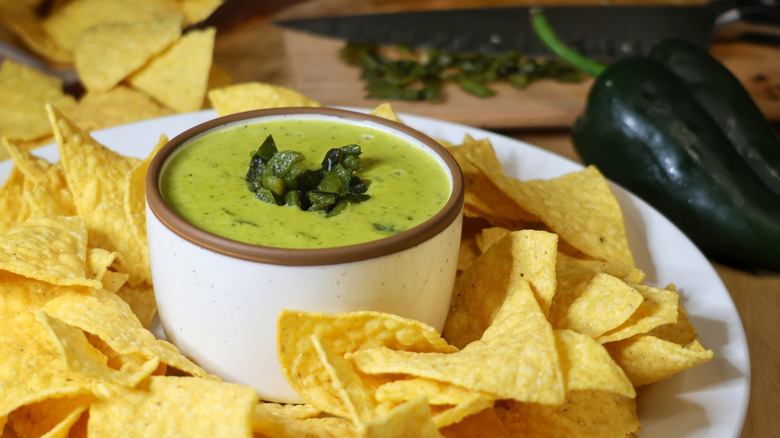 small bowl of poblano queso on a plate with tortilla chips and poblano peppers in the background