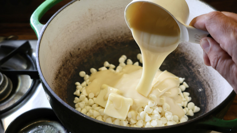 condensed milk pouring from measuring cup into pot of white chocolate chips and butter