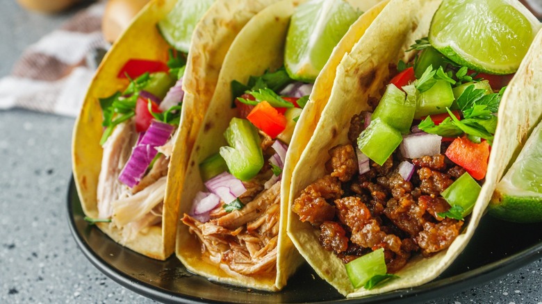 A trio of homemade tacos on a plate with different proteins as well as lime, cilantro, onion and tomato on top