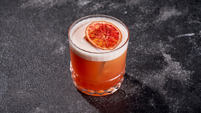 The drink is an appealing peachy-orange color, topped with a dense, white foam or froth, suggesting a sophisticated sour or egg white cocktail