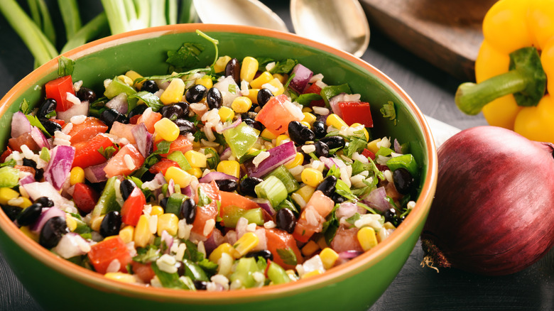 A green bowl of cowboy caviar sits beside a yellow bell pepper and a red onion, with two spoons resting in the background