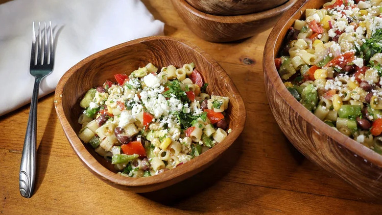 A serving of cowboy style pasta salad in a wooden bowl on a wooden table, with a fork resting beside it, next to a larger wooden bowl containing more of the pasta salad