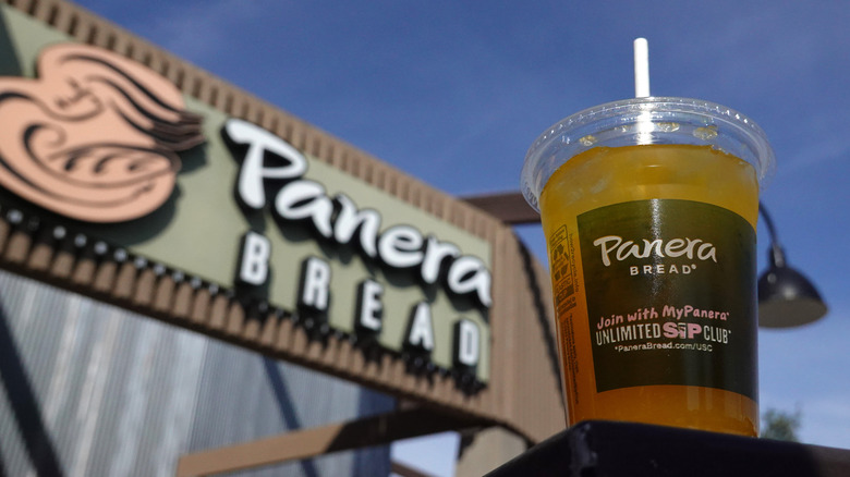 A Panera Bread branded plastic cup filled with Charged Lemonade and advertising the Unlimited Sip Cup stands in front of a Panera Bread location during daytime.