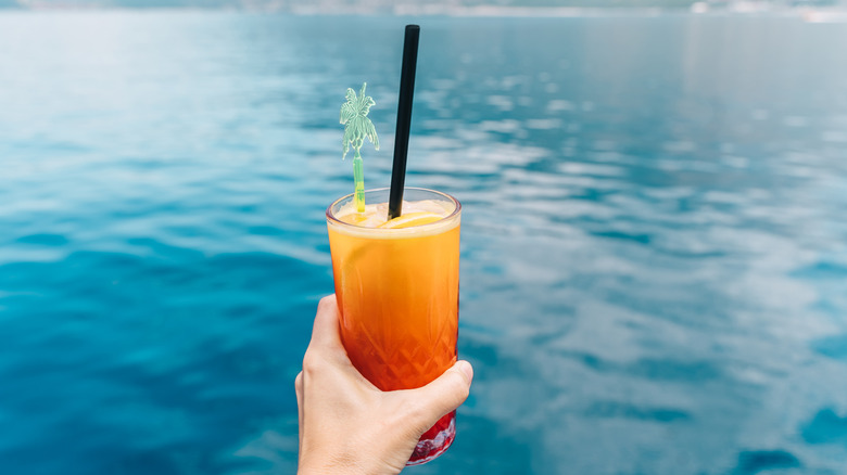 Person holding s*x on the beach cocktail in front of water background
