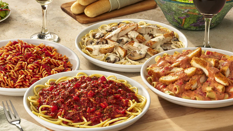 A variety of Olive Garden pasta dishes on a table with wine, salad, and breadsticks