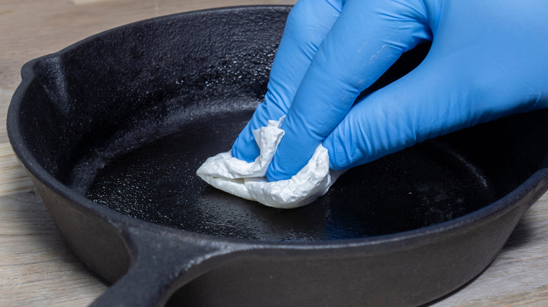 A blue nitrile-gloved hand seasons a cast iron pan with a paper towel