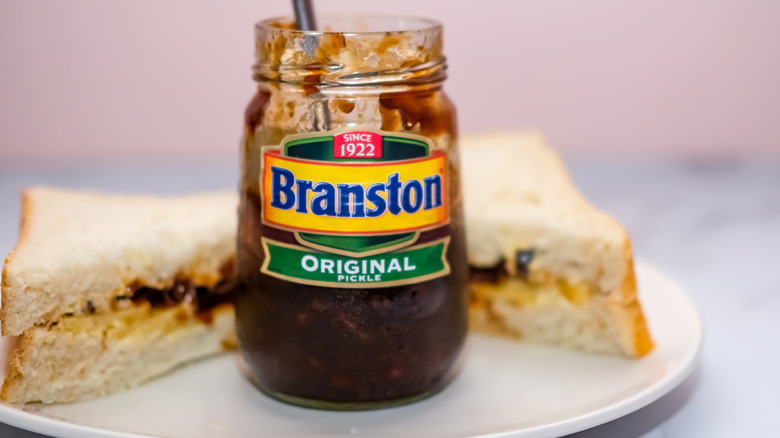 Jar of Branston Pickle on a white plate with a cheese and pickle sandwich