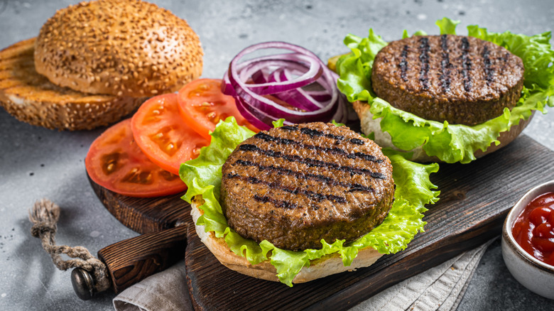 Vegan grilled patties, tomato, and onion in an open burger bun on a wooden serving board