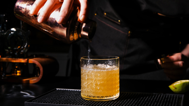 A bartender pouring a drink from a cocktail shaker into a glass, sitting beside a basket of limes
