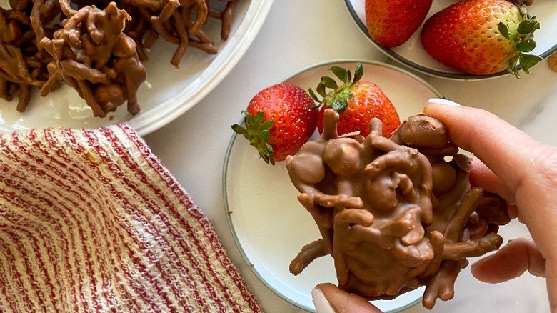 Hand holding chocolate peanut butter haystack cookie over table with small plates of strawberries and striped red and white cloth