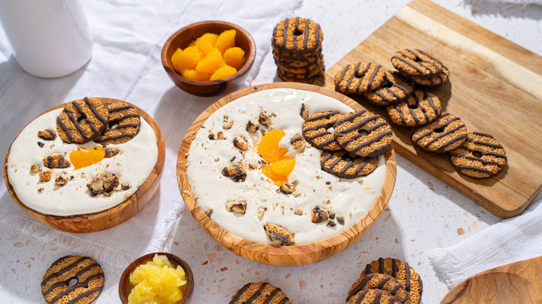 Two bowls of a white creamy substance are topped with whole and crushed chocolate striped cookies with more cookies and bowls of fruit on the table around them