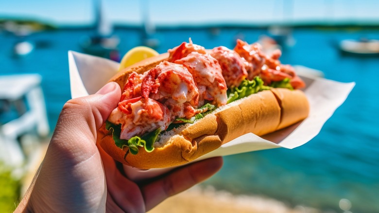 Hand holding a lobster roll in front of a New England coastline
