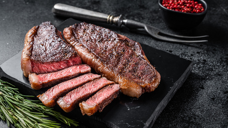 Ribeye medium rare on a black countertop with sprigs of rosemary and red peppercorns in background