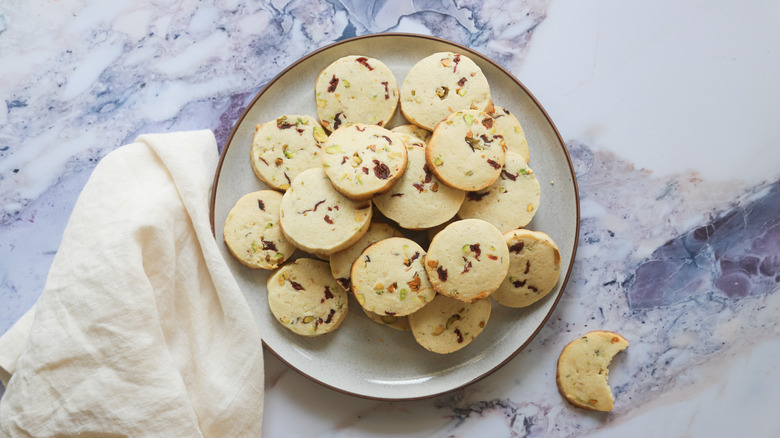 Pistachio cranberry cookies on a white plate beside a cloth napkin on a stone blue and white countertop