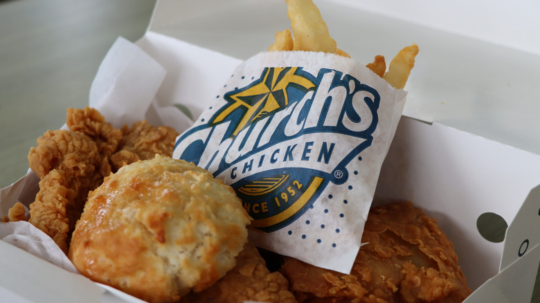 A white paper takeout box containing Church's Texas Chicken fried chicken, fries, and biscuits