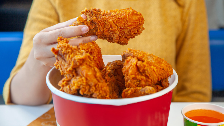 A person holding a fried chicken drumstick from a paper bucket of fried chicken