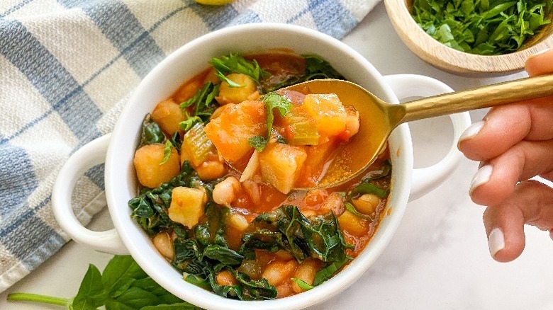 Hand holding spoon over bowl of potato, white bean, and kale stew