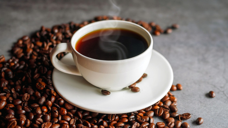 Black coffee in a white mug on a saucer, with whole coffee beans scattered next to it.