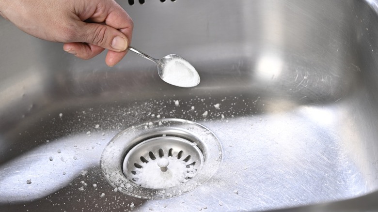 A hand dumping a spoonful of baking soda into an aluminum sink drain