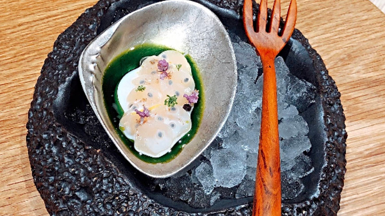 Scallop with basil oil on ice cubes in a metal bowl on a decorative black plate with an orange fork at Somni