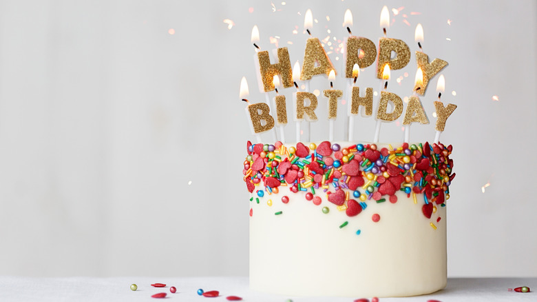 White cake decorate with colorful sprinkles and burning candles that spell out Happy Birthday.