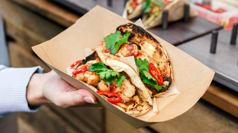 Hand holding a paper plate with two tacos, filled with meat, red peppers, and cilantro.