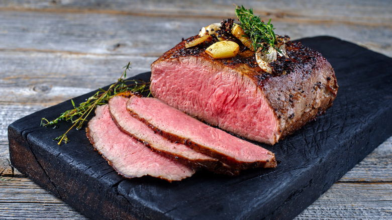 A thick piece of roast beef with three thin slices cut, served on a black wooden board.