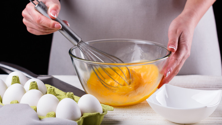 A woman whisks eggs in a glass bowl beside a carton of eggs