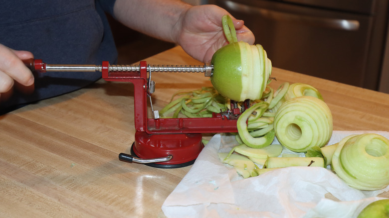 A red apple peeler sitting on a kitchen table as a man uses it to peel and slice green apples