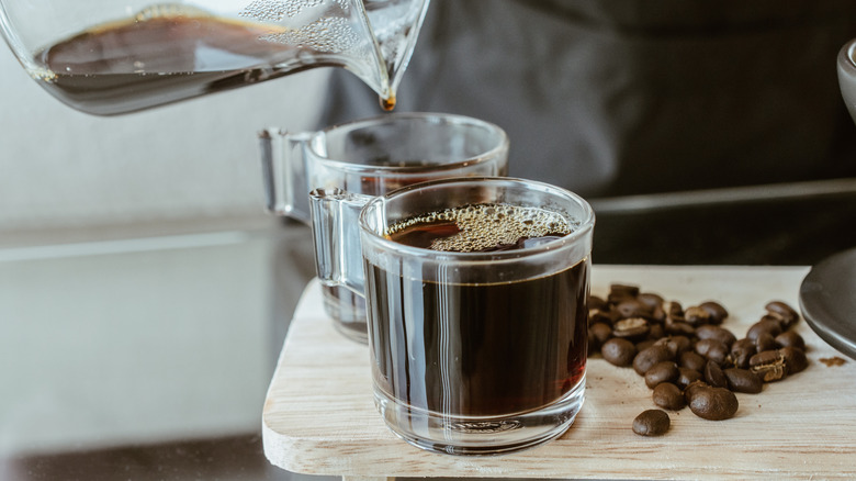 Someone pouring coffee from a pot into two steaming mugs, beside a pile of coffee beans