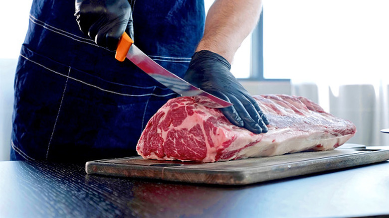 A butcher wearing an apron is cutting steak from a large portion of beef on a cutting board