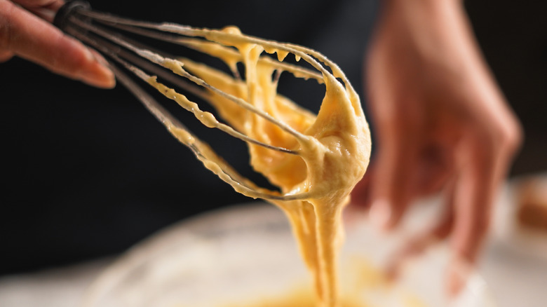 woman's hand lifting a whisk full of batter from a glass bowl, against a dark background