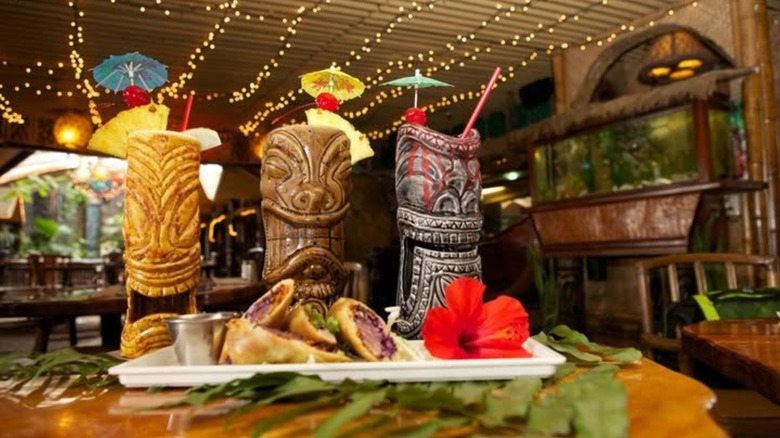 Three tiki mugs on a table with a plate of food against a blurred background