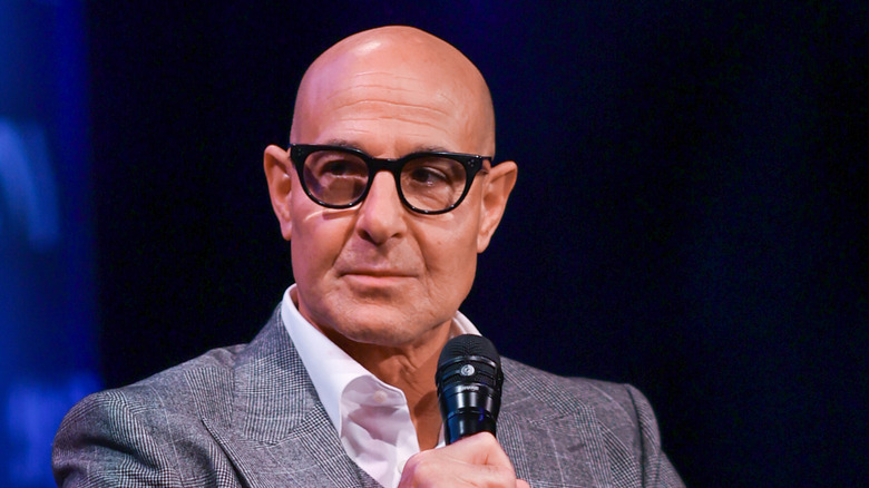 Stanley Tucci wearing dark-rimmed glasses in a white shirt and suit jacket, holding a microphone