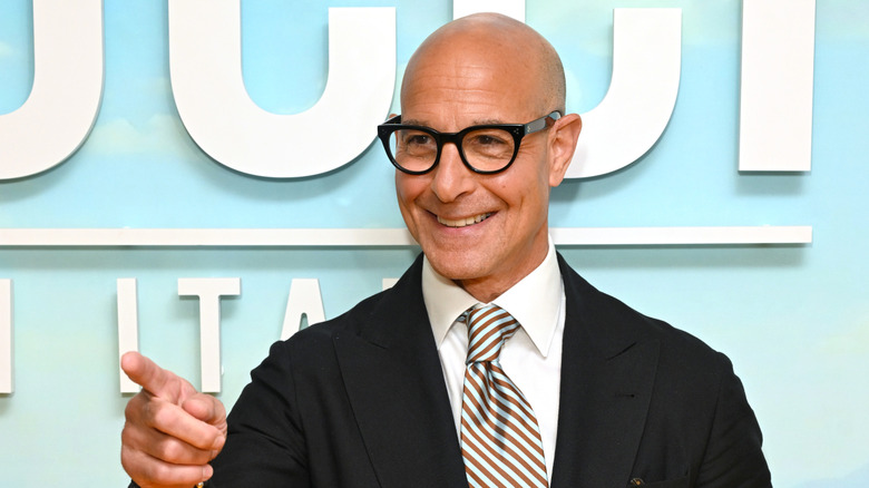 Stanley Tucci in a dark jacket with a striped tie, pointing to the left of the screen