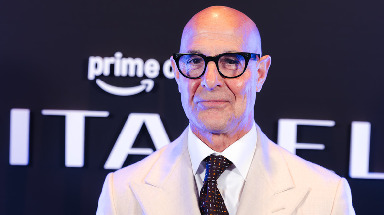 Stanley Tucci wearing a cream-colored suit and dark tie in front of an Amazon Prime backdrop