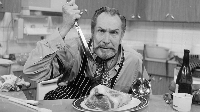 Vincent Price wearing a neckerchief and an apron, leaning over a cut of meat with a knife poised to stab it