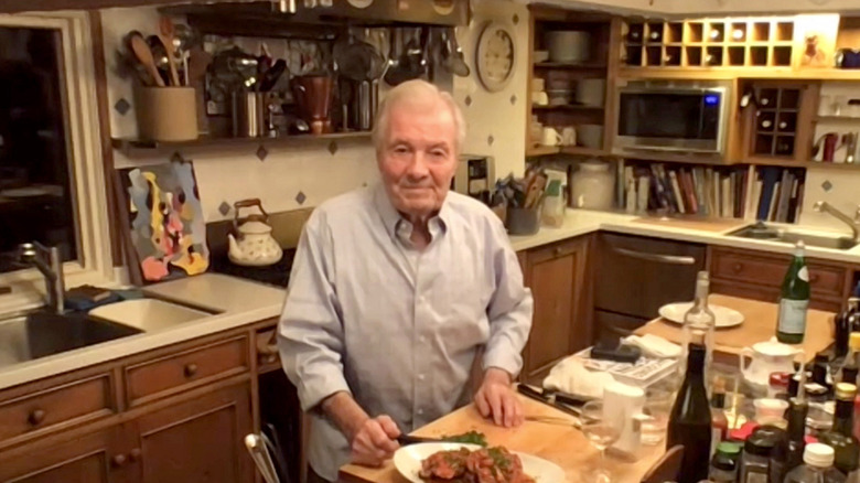 Jacques Pépin in a cluttered kitchen standing at a counter with a plate of food