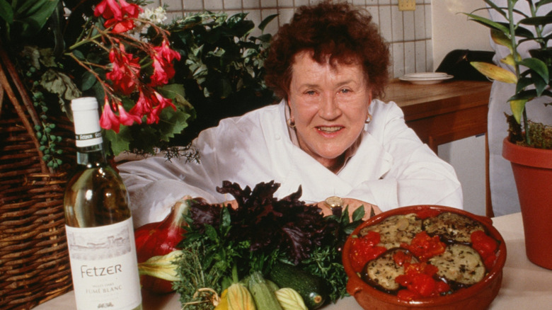 Photo of Julia Child leaning against a table filled with vegetables and flowers.