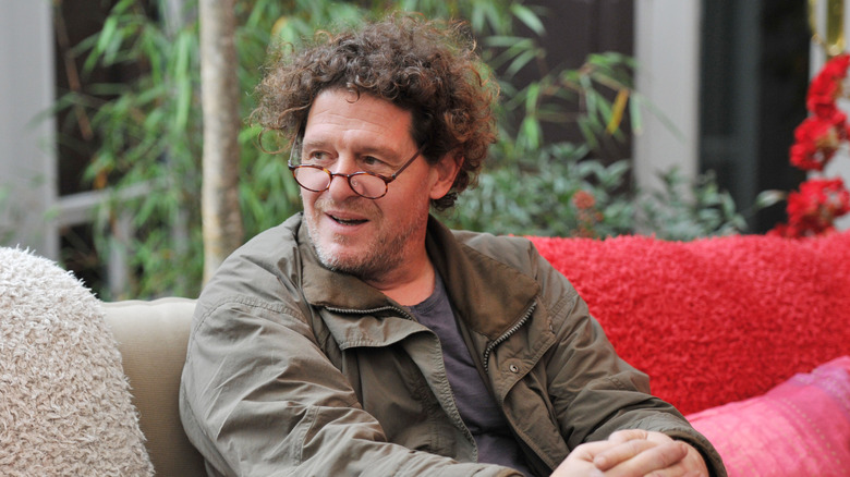 Marco Pierre White sitting on a couch, looking to the side, wearing a green jacket and glasses.