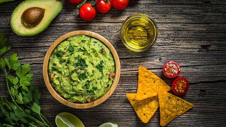 A bowl of guacamole dip, avocado, tomatoes, and chips