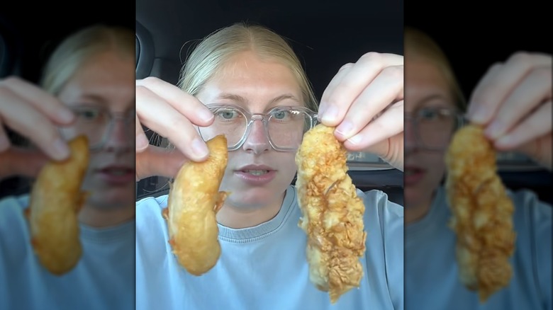 A woman holding two chicken fingers up to demonstrate the difference between their batter coating.