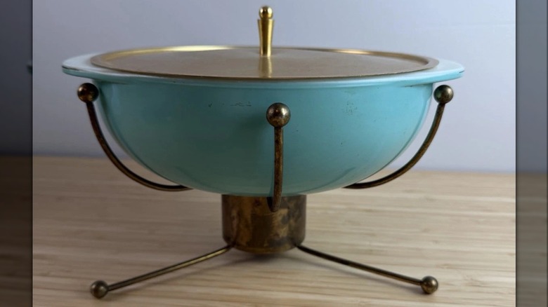 A turquoise and brass Pyrex Super DeLuxe Casserole dish on a warming stand