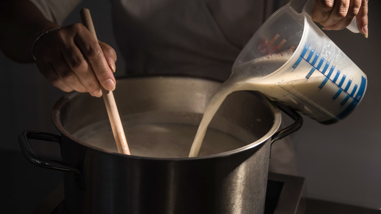 person pouring milk into a metal pot on the stove while stirring using a wooden spoon with their other hand